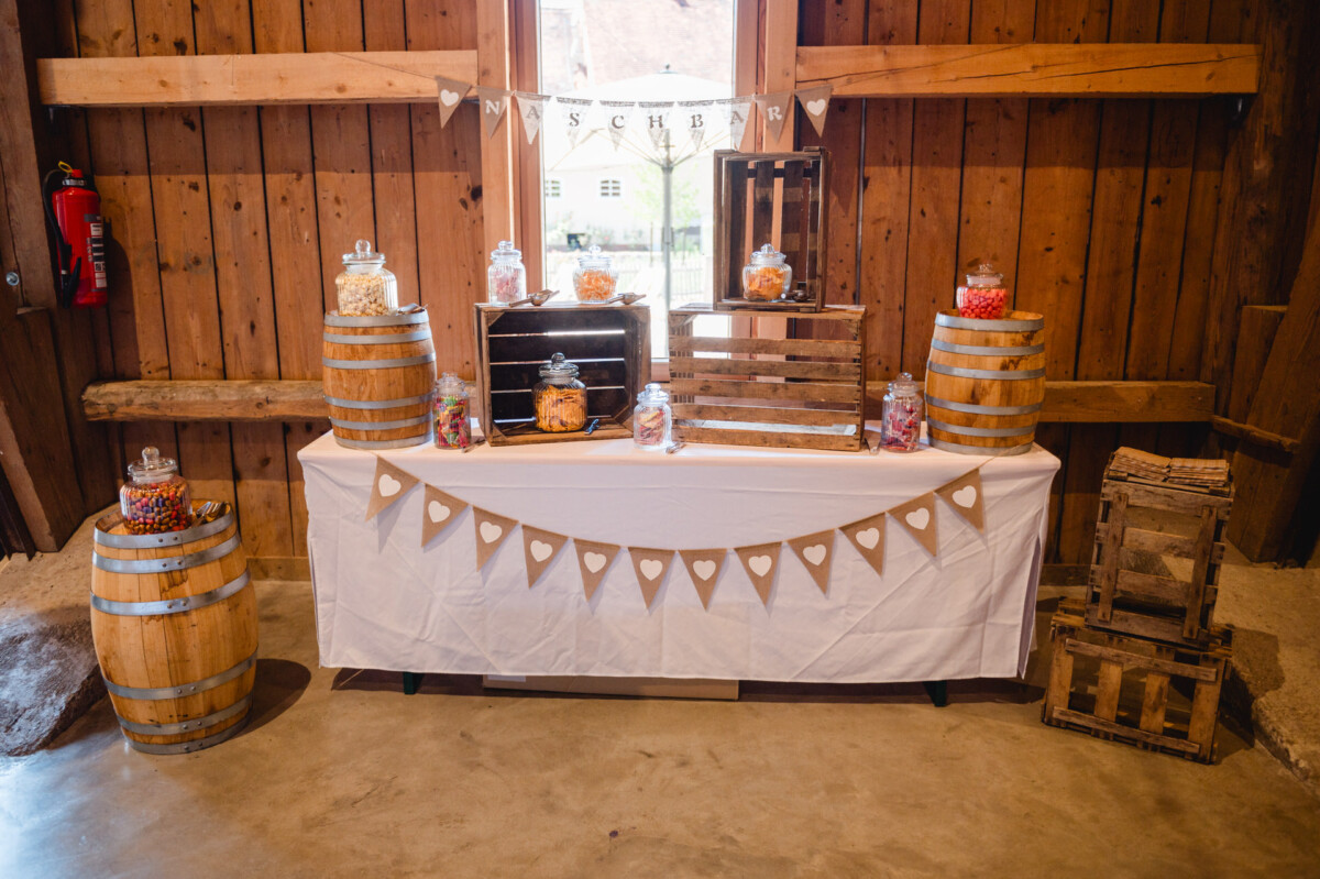 Eine rustikale Süßigkeitenauslage mit Gläsern mit verschiedenen Süßigkeiten auf Holzfässern und Kisten. Eine weiße Tischdecke und eine Herzgirlande schmücken den Tisch.