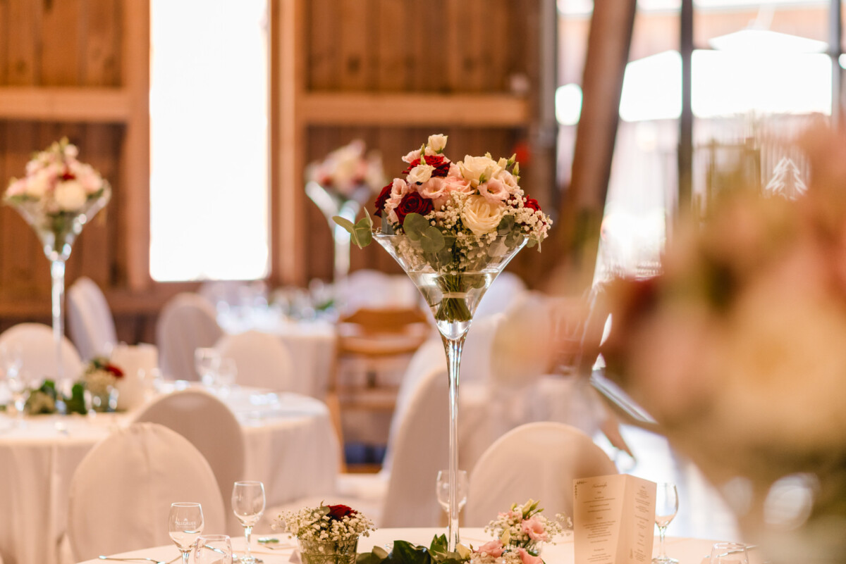 Eleganter Hochzeitsempfang mit runden Tischen, weißen Tischdecken und großen Blumengestecken mit roten und rosa Rosen.