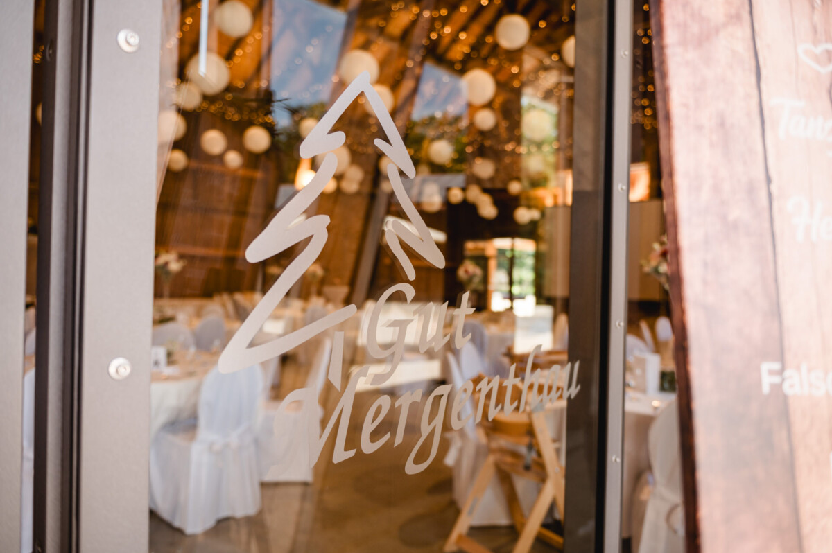 Glastür mit einem mattierten Baumlogo und dem Text „Gut Hergertshau“ bietet Blick auf einen dekorierten Innenraum mit Tischen, Stühlen und Hängelampen.