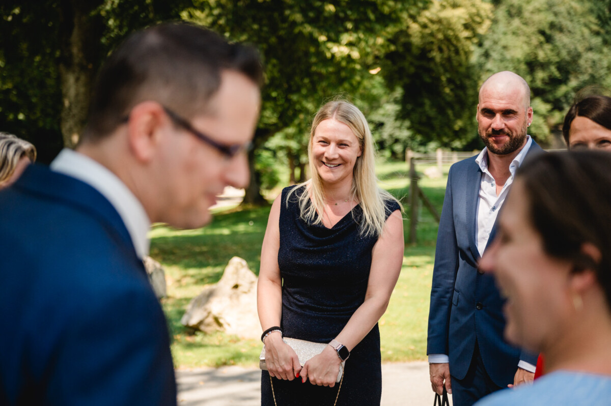 Eine Gruppe von Menschen in formeller Kleidung steht draußen, lächelt und unterhält sich. Im Hintergrund sind Bäume und Grün zu sehen.