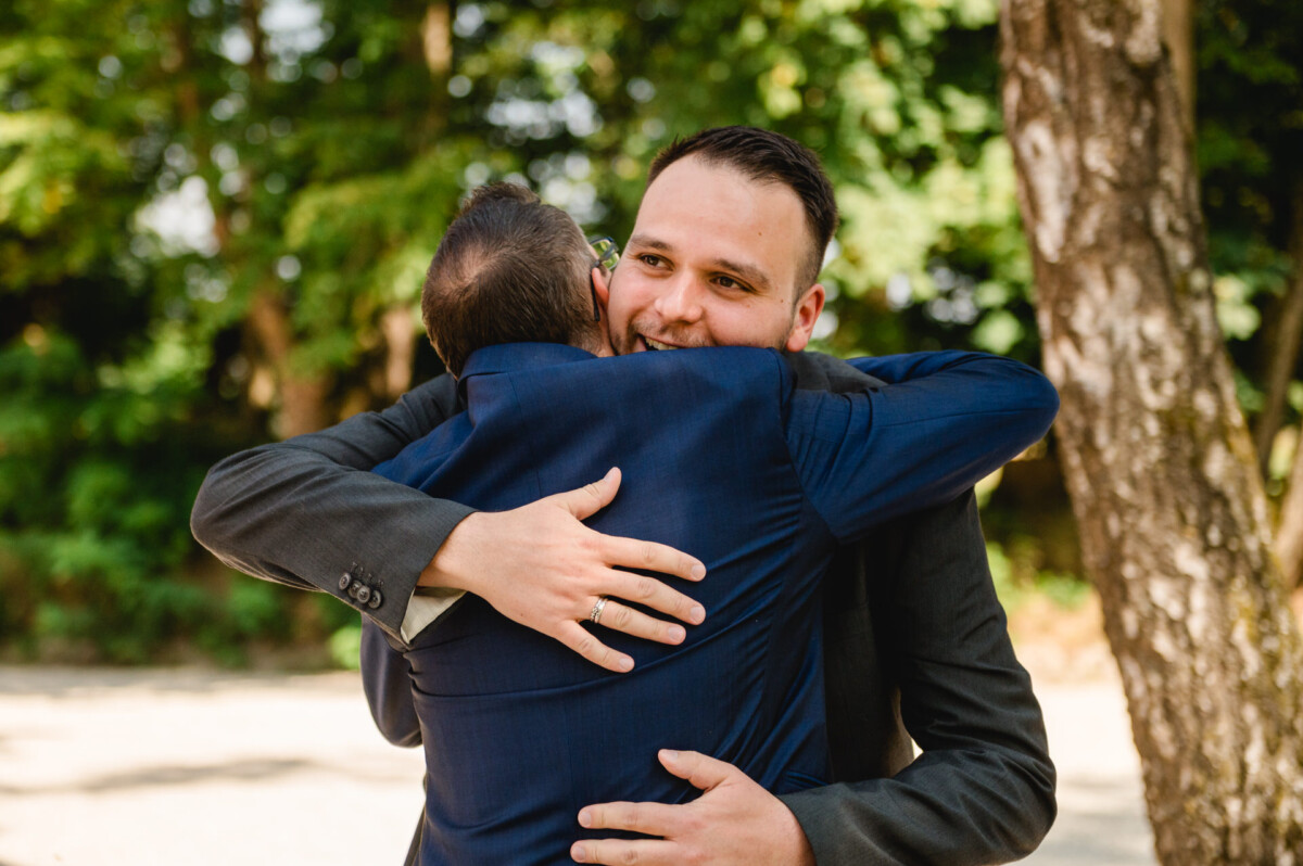 Zwei Männer in Anzügen umarmen sich im Freien neben Bäumen.
