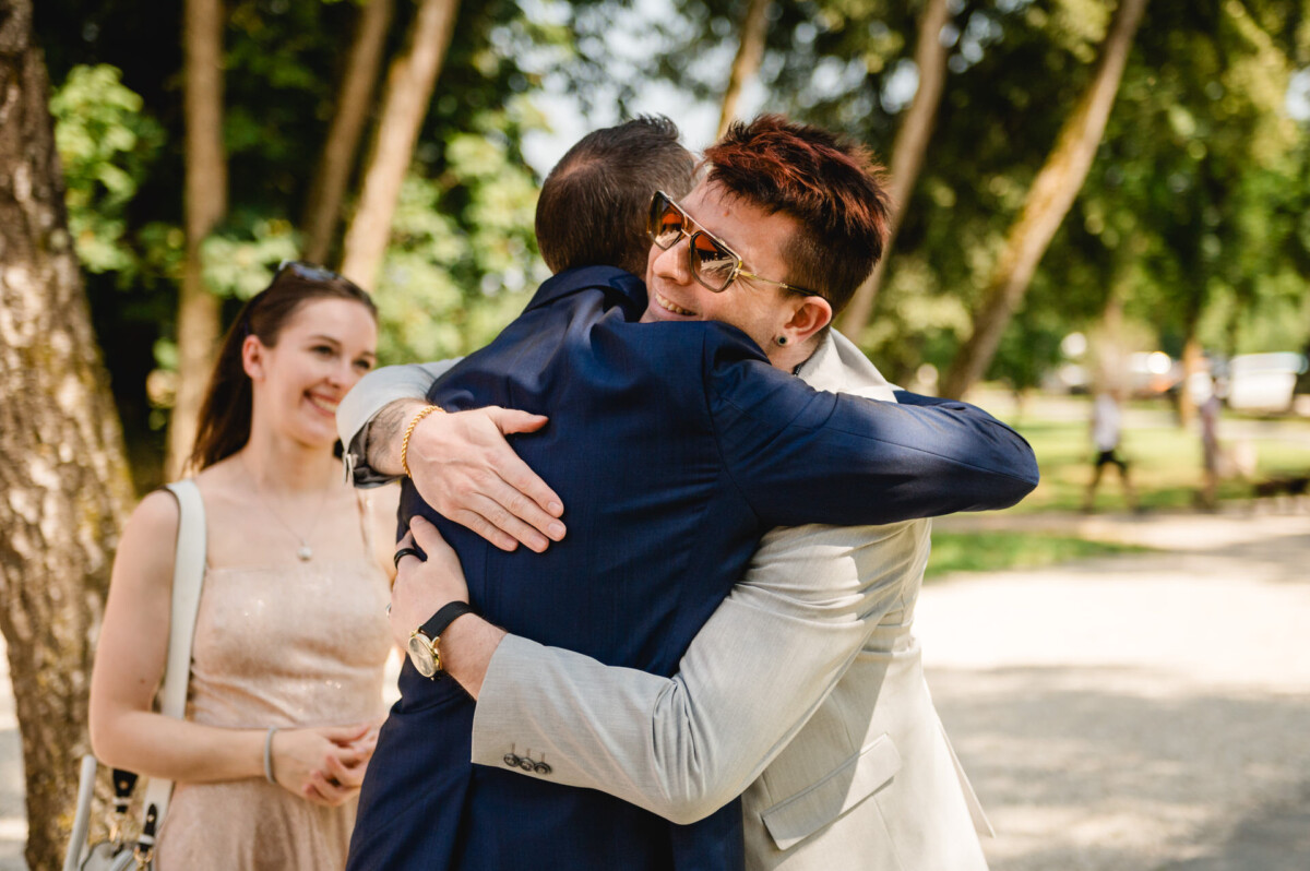 Zwei Männer in Anzügen umarmen sich herzlich im Freien, daneben steht eine lächelnde Frau. Im Hintergrund sind Bäume und Sonnenlicht zu sehen.