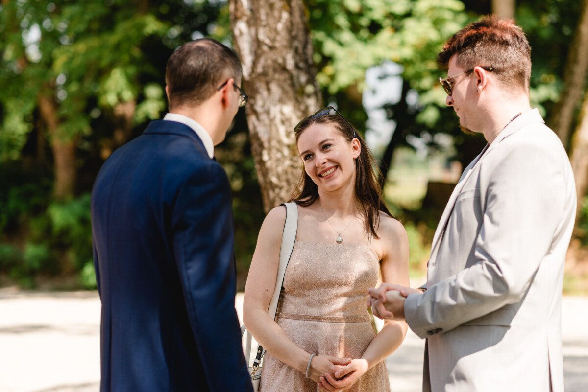 Drei Personen unterhalten sich im Freien, im Hintergrund Bäume. Die Frau lächelt, während zwei Männer in Anzügen daneben stehen.