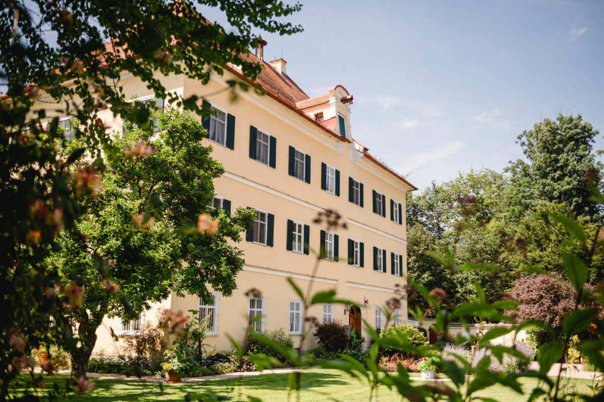 Ein großes gelbes Gebäude mit grünen Fensterläden und einem roten Dach, umgeben von Bäumen und Pflanzen, unter einem klaren blauen Himmel.