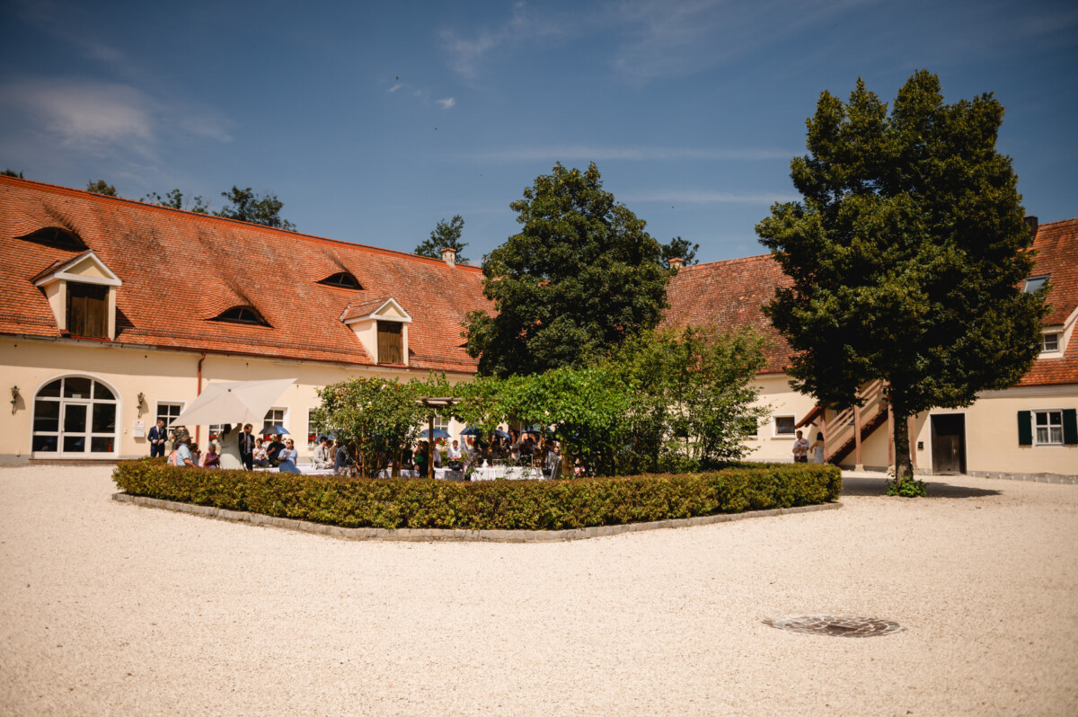 Historischer Innenhof mit Gebäuden mit orangefarbenen Dächern, einem gepflegten Garten und einer Menschenansammlung unter einem Zelt an einem sonnigen Tag.