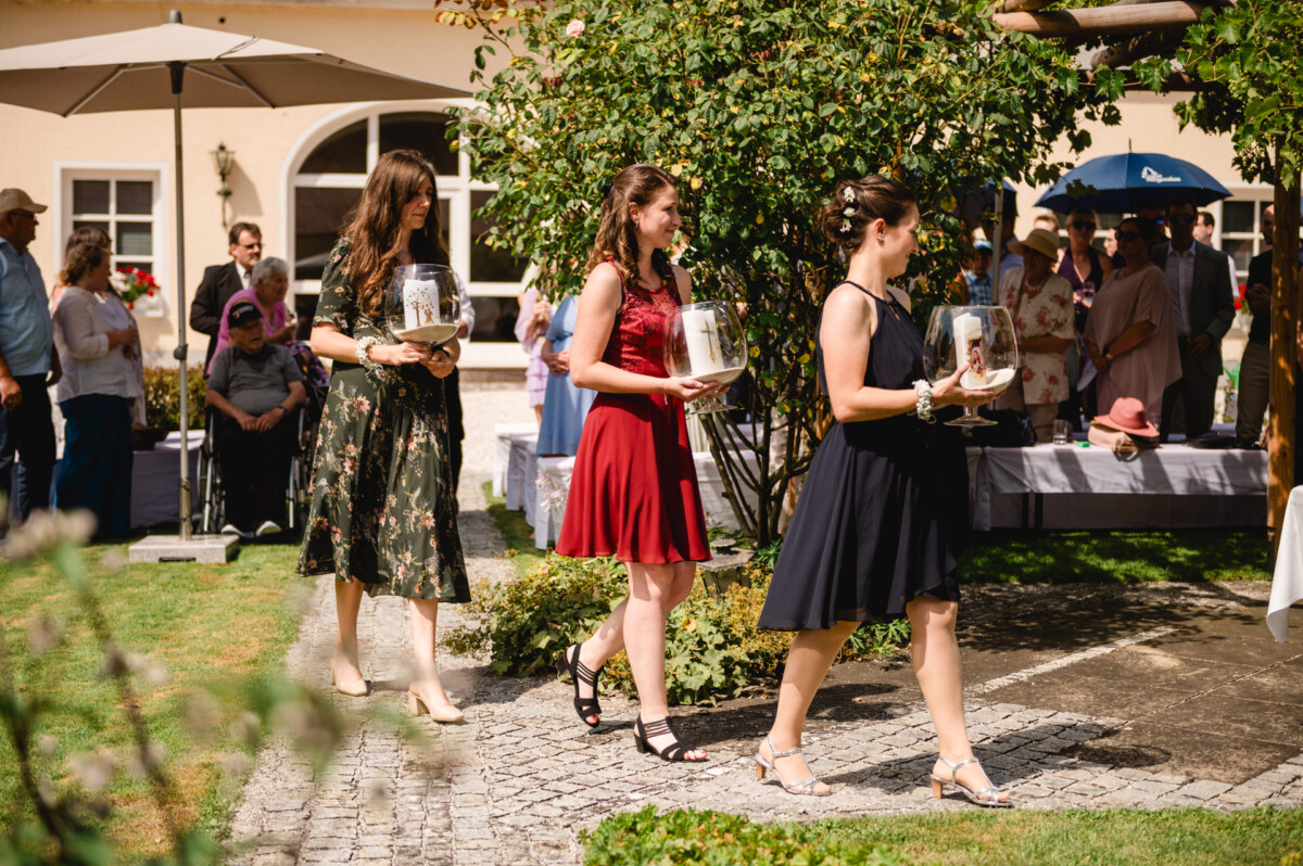 Drei Frauen in Kleidern gehen durch einen Garten. Jede trägt ein Glas mit einer Kerze. Im Hintergrund sitzen Menschen.
