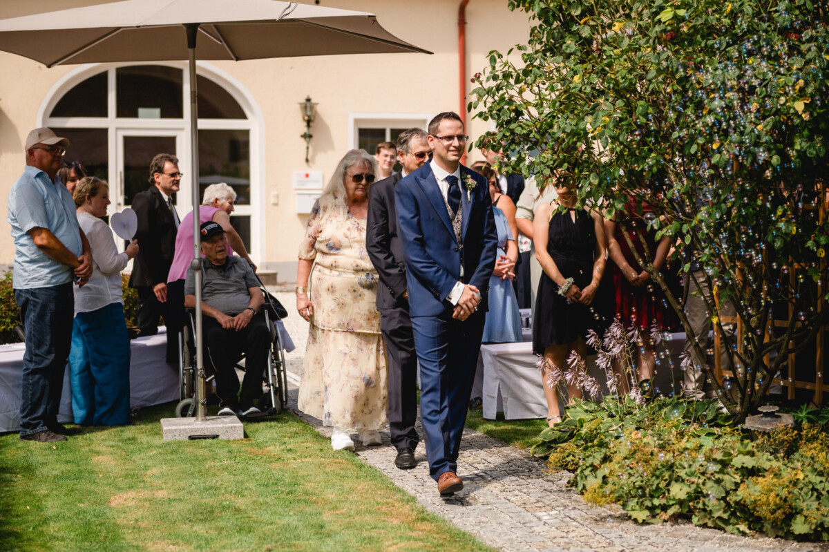 Eine Gruppe von Menschen, darunter ein Mann im Anzug, versammelt sich draußen unter einem Sonnenschirm, in der Nähe steht ein Rollstuhl und es gibt Grünflächen.