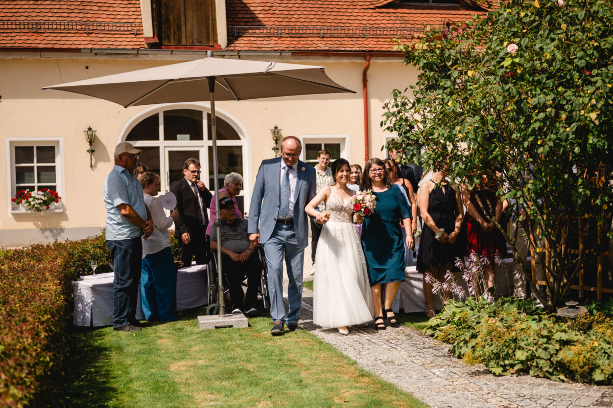 Eine Braut im weißen Kleid geht Arm in Arm mit einem Mann im Anzug einen Gartenweg entlang. Zuschauer stehen in der Nähe unter einem großen Sonnenschirm vor einem gelben Gebäude.