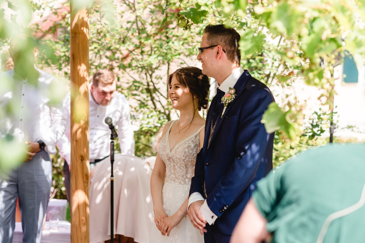 Eine Braut und ein Bräutigam lächeln während einer Hochzeitszeremonie im Freien, mit Grün im Hintergrund und Gästen in der Nähe.