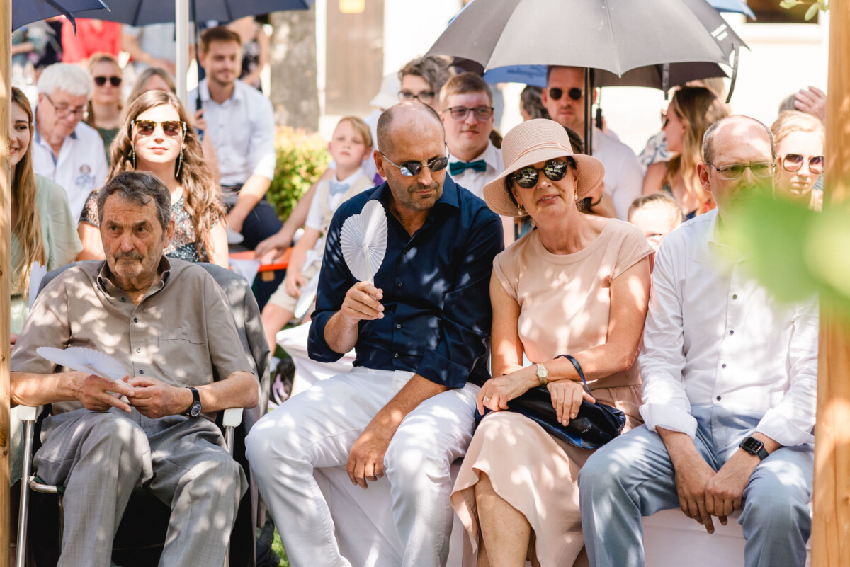 Eine Gruppe von Menschen sitzt im Freien, einige halten Regenschirme und Fächer in den Händen. Sie tragen Sommerkleidung und wirken entspannt, wie sie in einer sonnigen Umgebung auf Klappstühlen sitzen.
