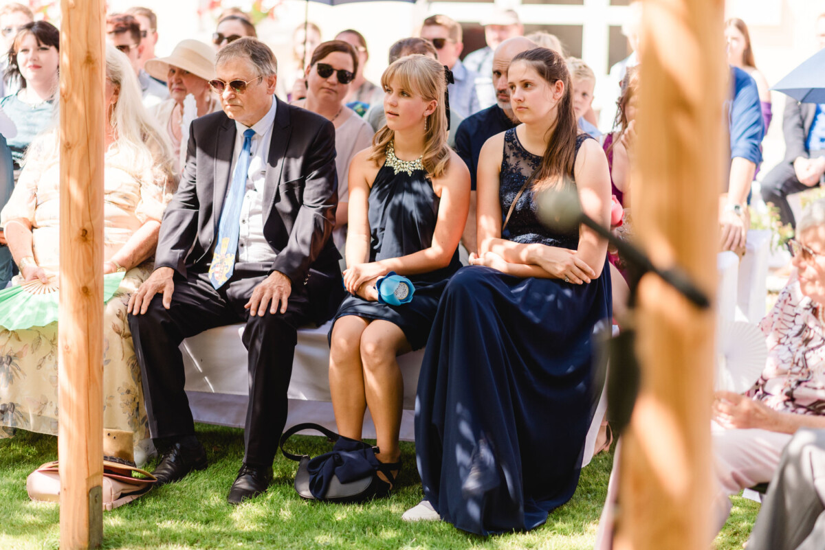 Menschen sitzen bei einer formellen Veranstaltung im Freien, die Männer in Anzügen und die Frauen in Kleidern, unter einem sonnigen Himmel.