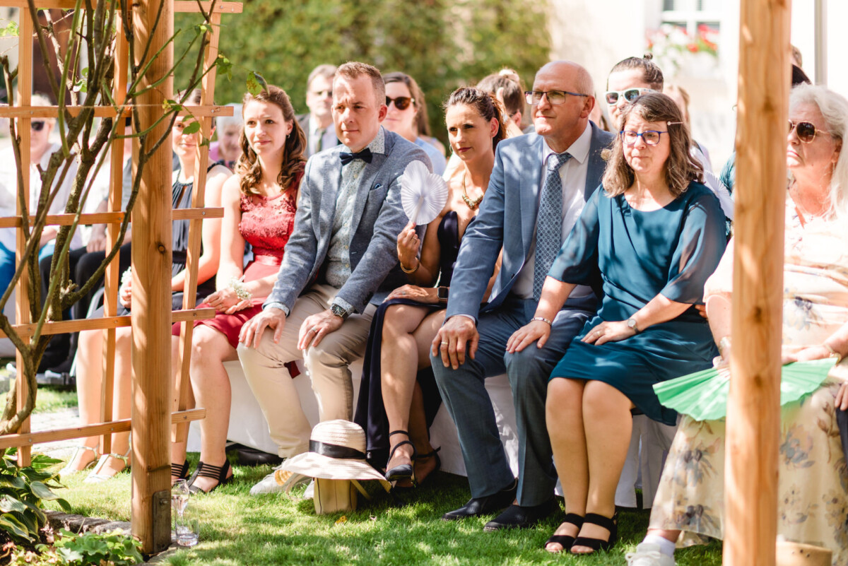 Eine Gruppe von Menschen sitzt bei einer Veranstaltung im Freien auf Bänken, einige halten Programme oder Ventilatoren in den Händen.