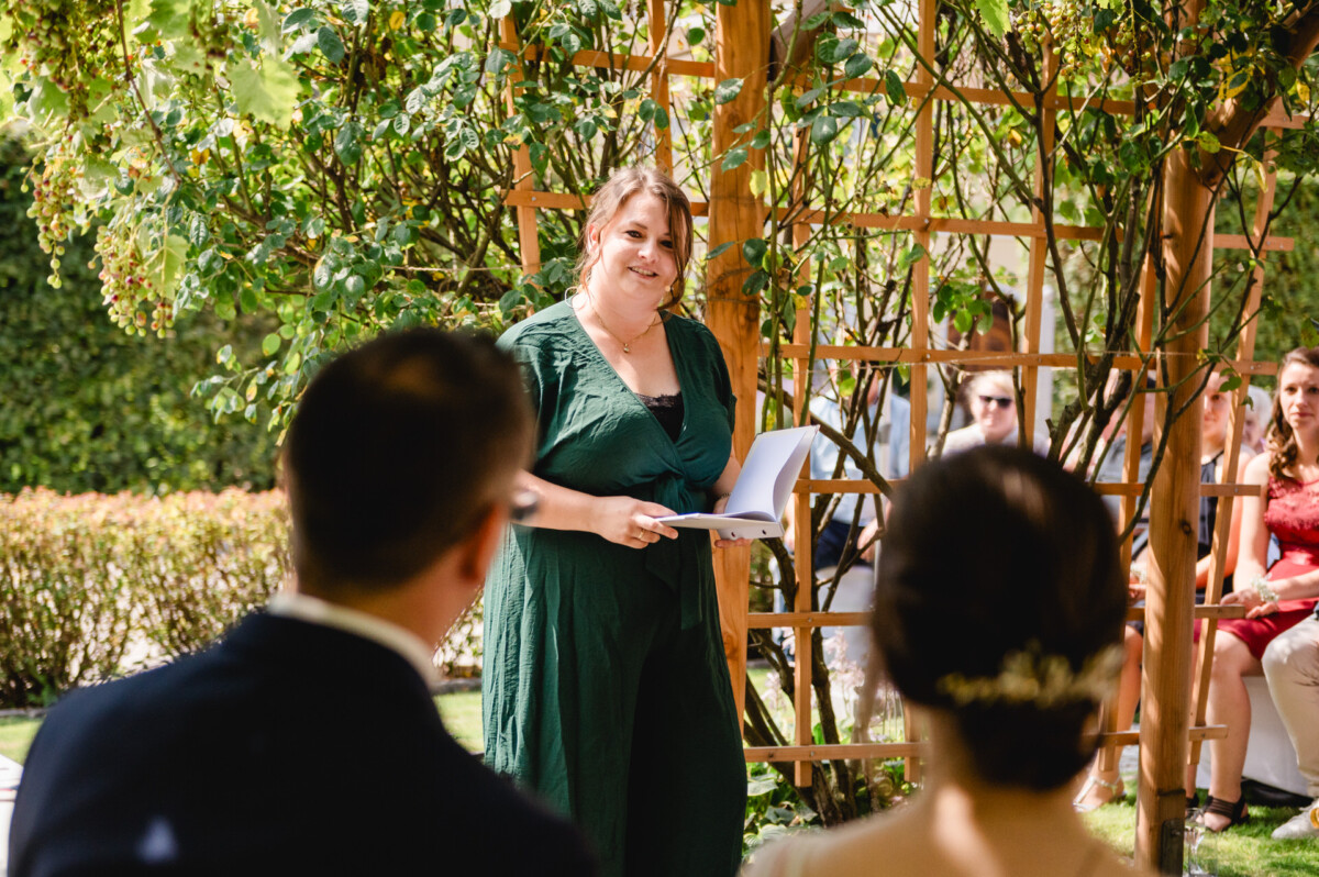 Eine Frau in einem grünen Kleid steht unter einer Holzlaube, hält Papiere in der Hand und lächelt ein sitzendes Paar an. Im Hintergrund sind mehrere Gäste zu sehen.
