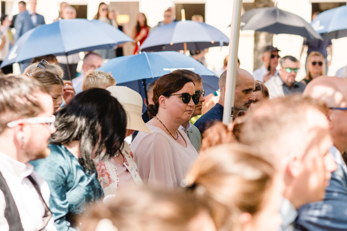 Eine Menschenmenge steht im Freien, viele halten blaue Regenschirme in der Hand. Einige tragen Sonnenbrillen. Die Umgebung scheint sonnig.
