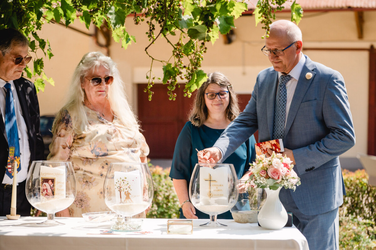 Ein Mann im Anzug interagiert mit einer Ausstellung großer Glasschalen auf einem Tisch, begleitet von drei Personen unter belaubten Zweigen im Freien.