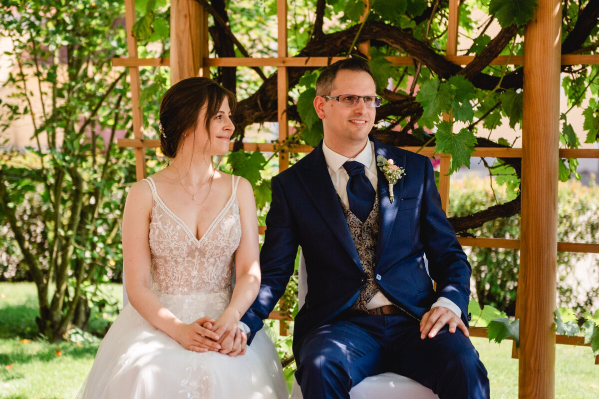 Ein Paar sitzt im Hochzeitskleid unter einer Pergola in einem Garten. Die Braut trägt ein weißes Kleid und der Bräutigam einen marineblauen Anzug.