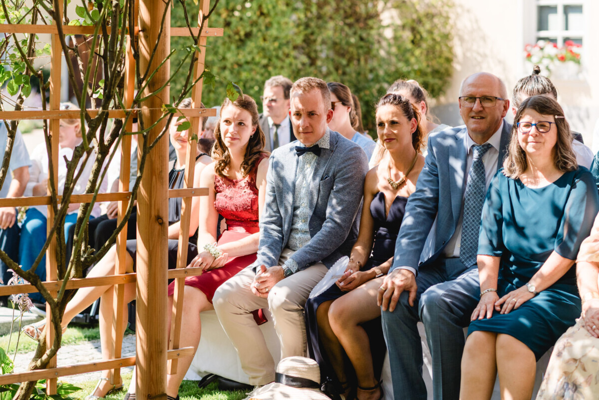 Gäste sitzen während einer Versammlung tagsüber im Freien, mit einer Holzpergola und Grünpflanzen im Vordergrund.