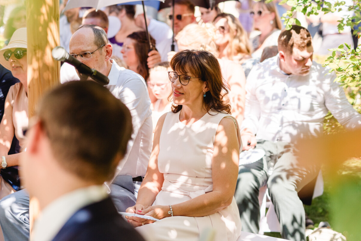 Eine Frau in einem weißen Kleid sitzt im Freien inmitten einer Menschenmenge. Einige halten Regenschirme in den Händen. Sonnenlicht fällt durch die Bäume und wirft Schatten.
