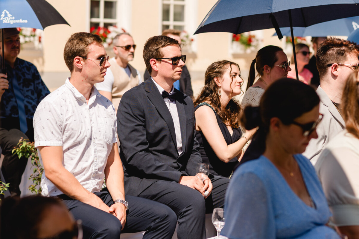 Eine Gruppe von Menschen sitzt bei einer Veranstaltung im Freien, einige halten Sonnenschirme in der Hand, um Schatten zu spenden. Sie scheinen aufmerksam zuzuschauen oder zuzuhören. Die Umgebung ist sonnig.