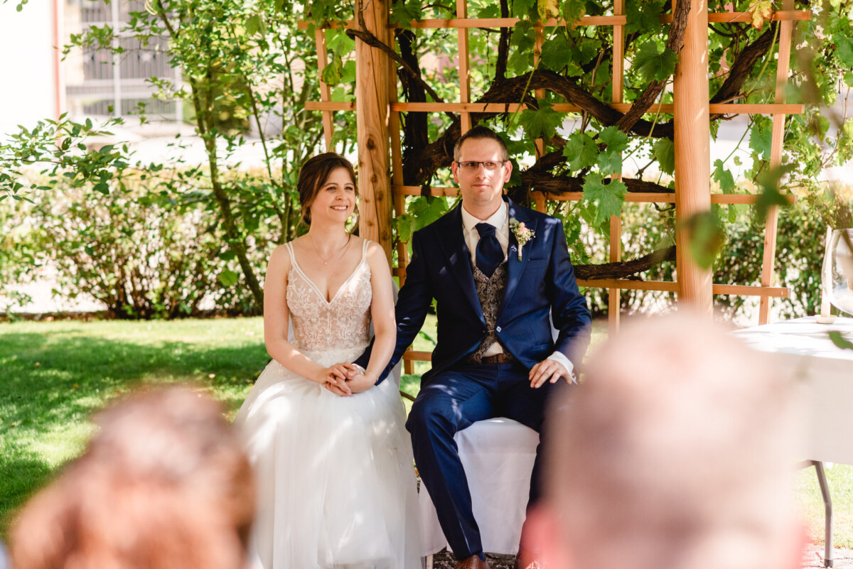 Braut und Bräutigam sitzen während einer Hochzeitszeremonie zusammen unter einer hölzernen Pergola in einer Gartenumgebung.