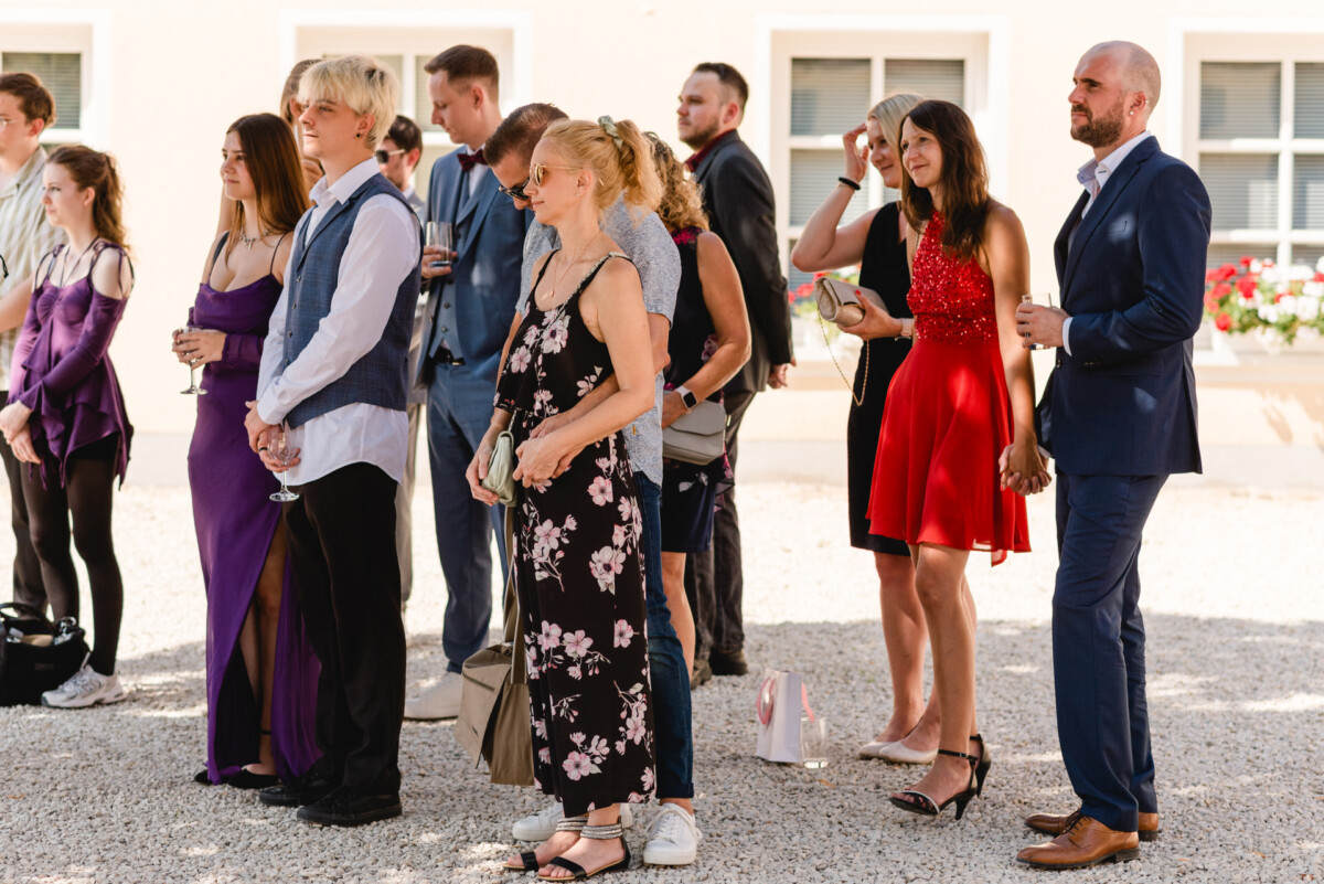 Eine Gruppe von Menschen in formeller Kleidung steht im Freien und beobachtet eine Veranstaltung. Einige halten Getränke in den Händen und man sieht verschiedene bunte Kleider und Anzüge.