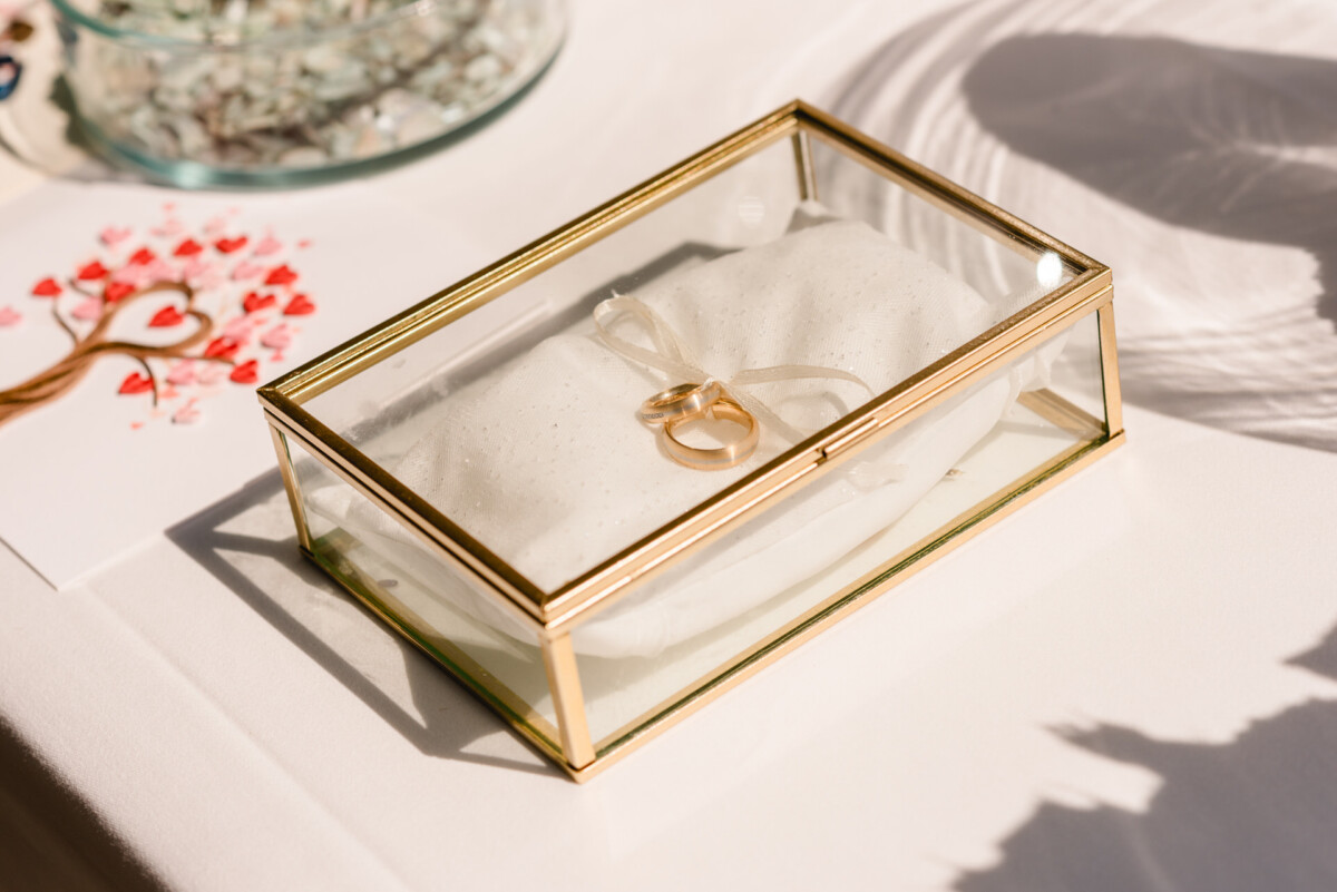 Zwei goldene Ringe auf einem weißen Kissen in einer Glasbox mit goldenem Rahmen, auf einem Tisch platziert.