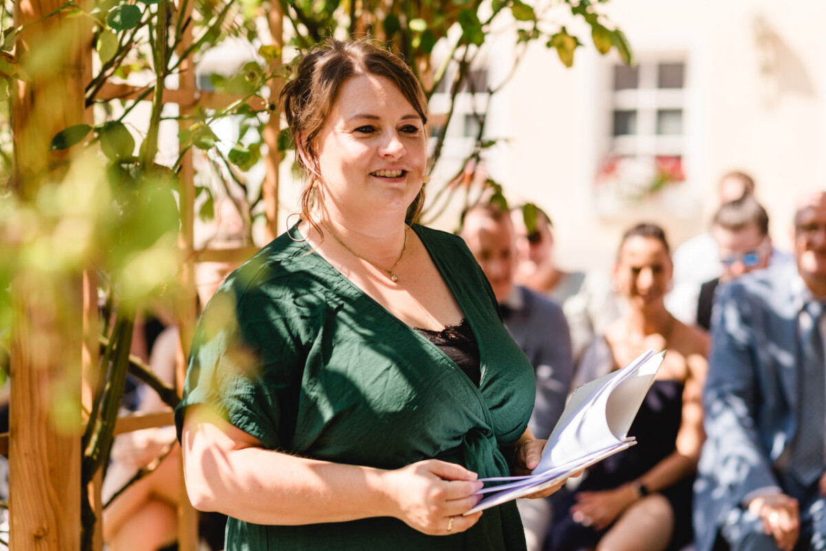 Eine Frau in einem grünen Kleid hält an einem sonnigen Tag im Freien eine Rede mit einem kleinen Publikum, das hinter ihr sitzt.