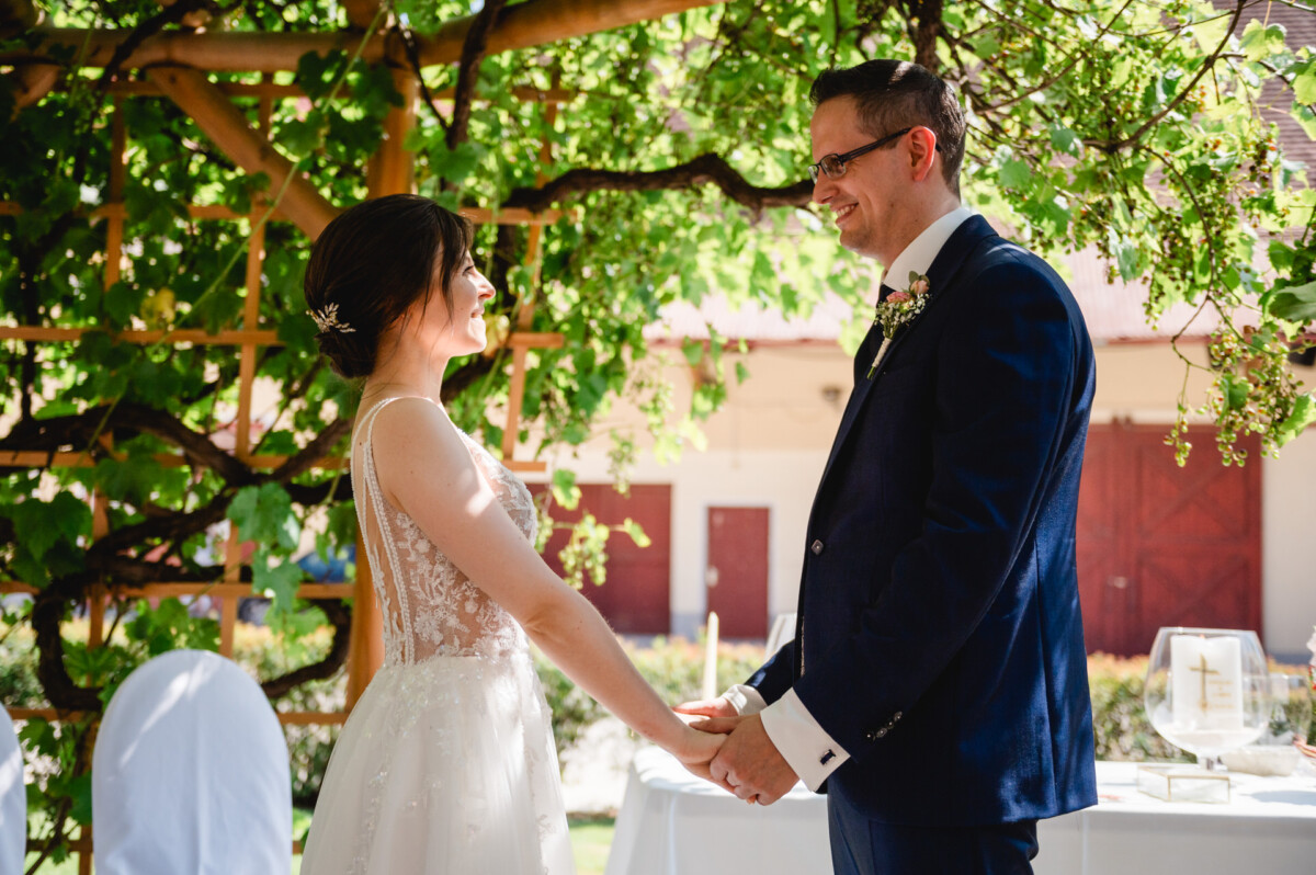 Eine Braut und ein Bräutigam halten im Freien unter einem Blätterdach Händchen und lächeln sich an.