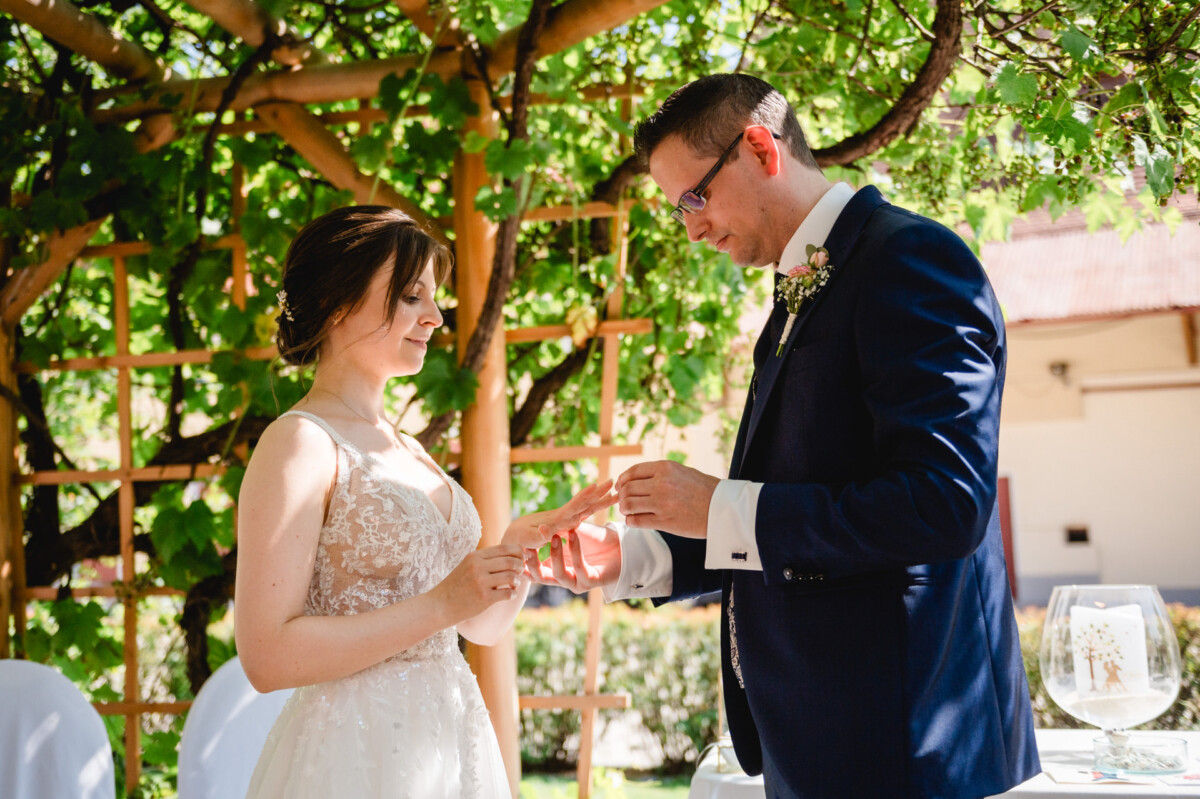 Ein Paar steht unter einem Spalier und tauscht Ringe. Die Frau trägt ein weißes Kleid und der Mann einen marineblauen Anzug. Es scheint eine Hochzeitszeremonie im Freien zu sein.