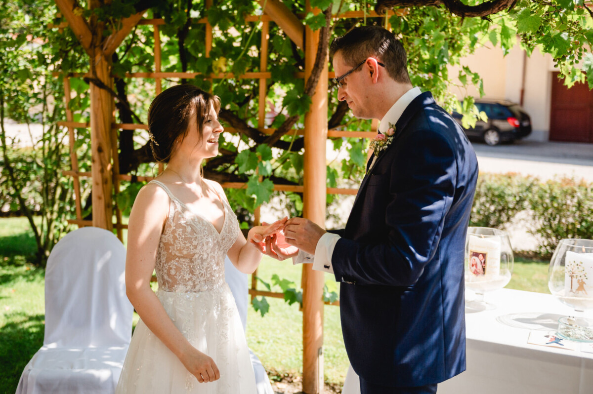 Ein Paar tauscht bei einer Hochzeitszeremonie im Freien unter einer weinbewachsenen Laube Ringe aus.