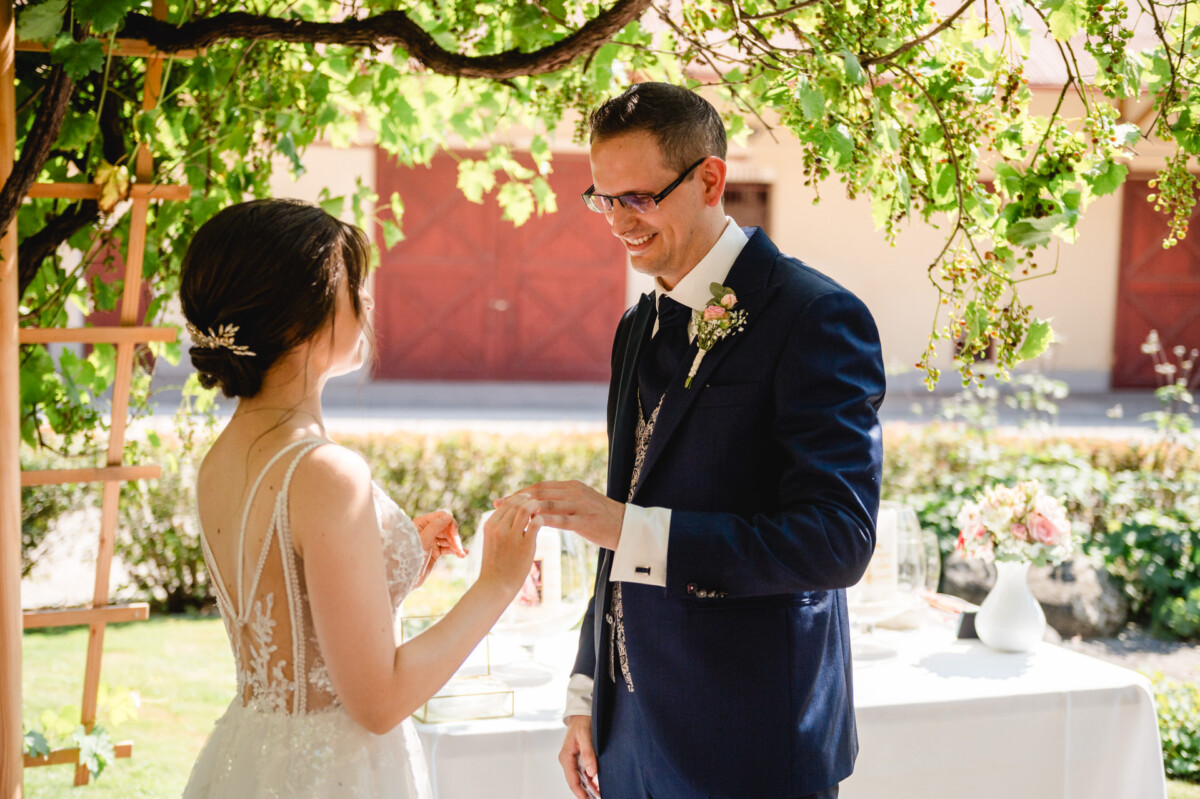 Braut und Bräutigam tauschen während einer Hochzeitszeremonie im Freien unter einem weinbewachsenen Torbogen die Ringe aus.