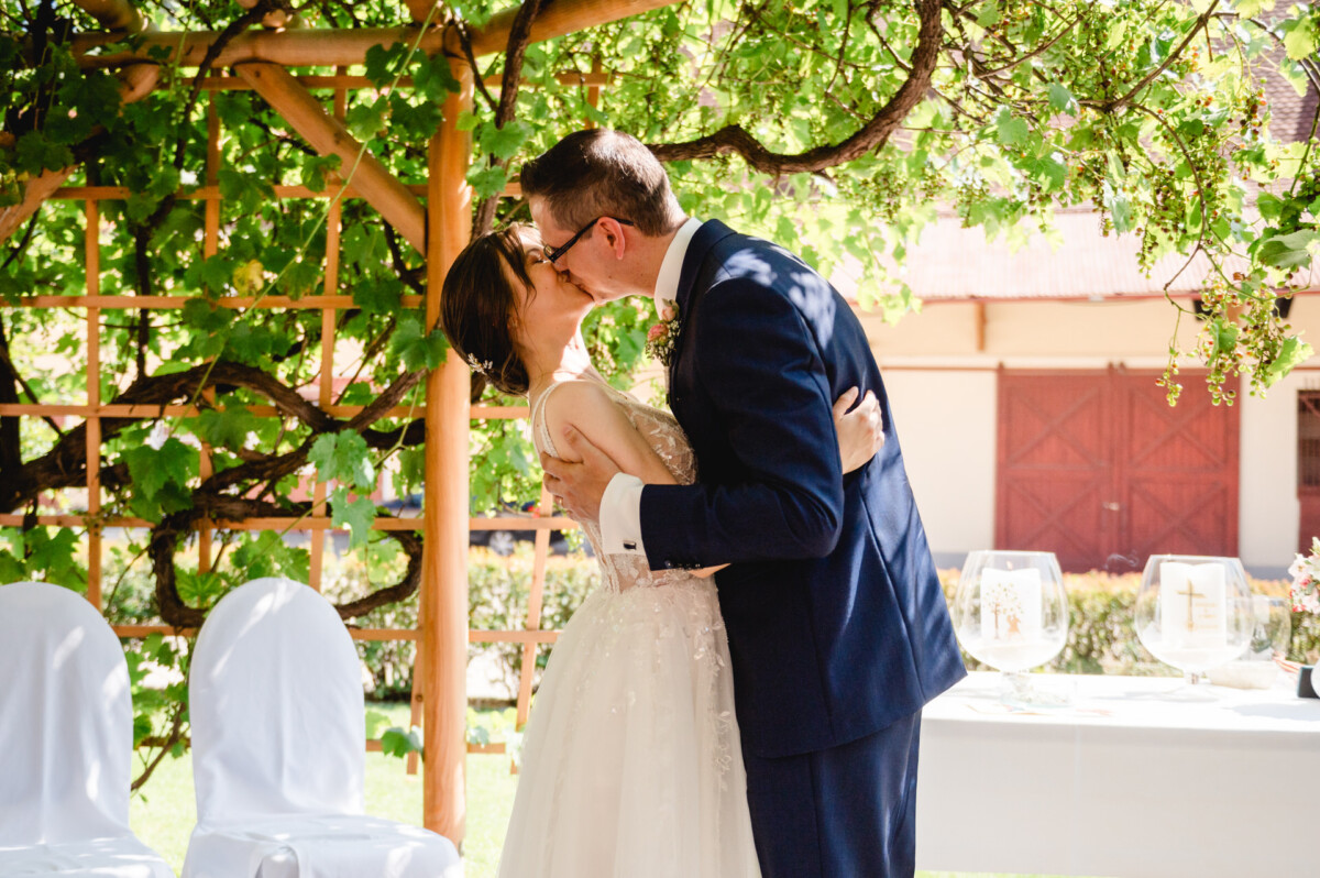Braut und Bräutigam küssen sich unter einer mit Grünpflanzen behangenen Pergola, neben einem mit Gläsern gedeckten Tisch und mit weißen Tüchern bedeckten Stühlen.