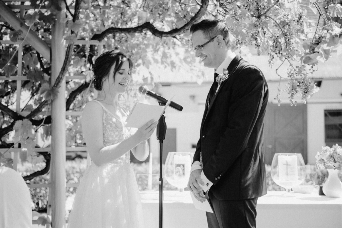 Braut und Bräutigam geben sich an einem sonnigen Tag im Freien das Ja-Wort. Die Braut liest aus einer Zeitung vor, während der Bräutigam zuhört und lächelt.