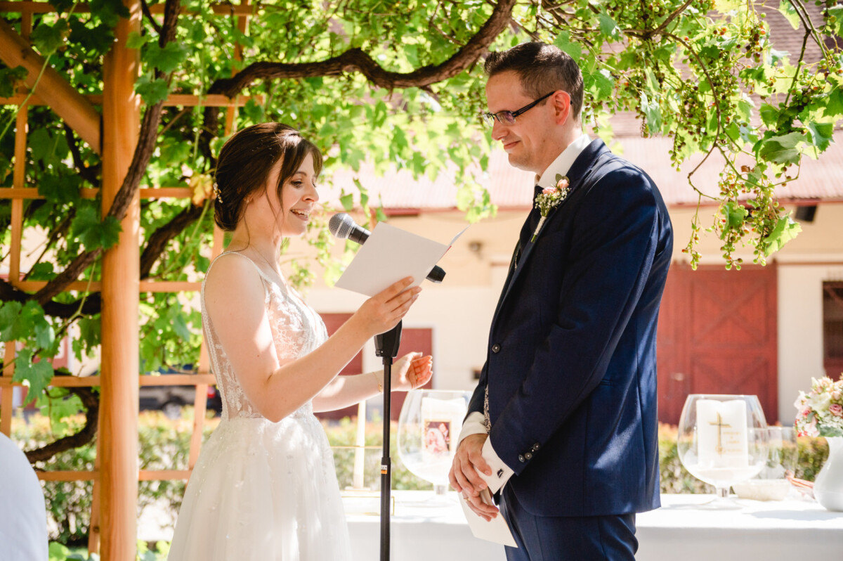 Braut und Bräutigam stehen während einer Hochzeitszeremonie im Freien im Grünen. Die Braut liest aus einer Zeitung, während der Bräutigam zuhört. Beide lächeln.