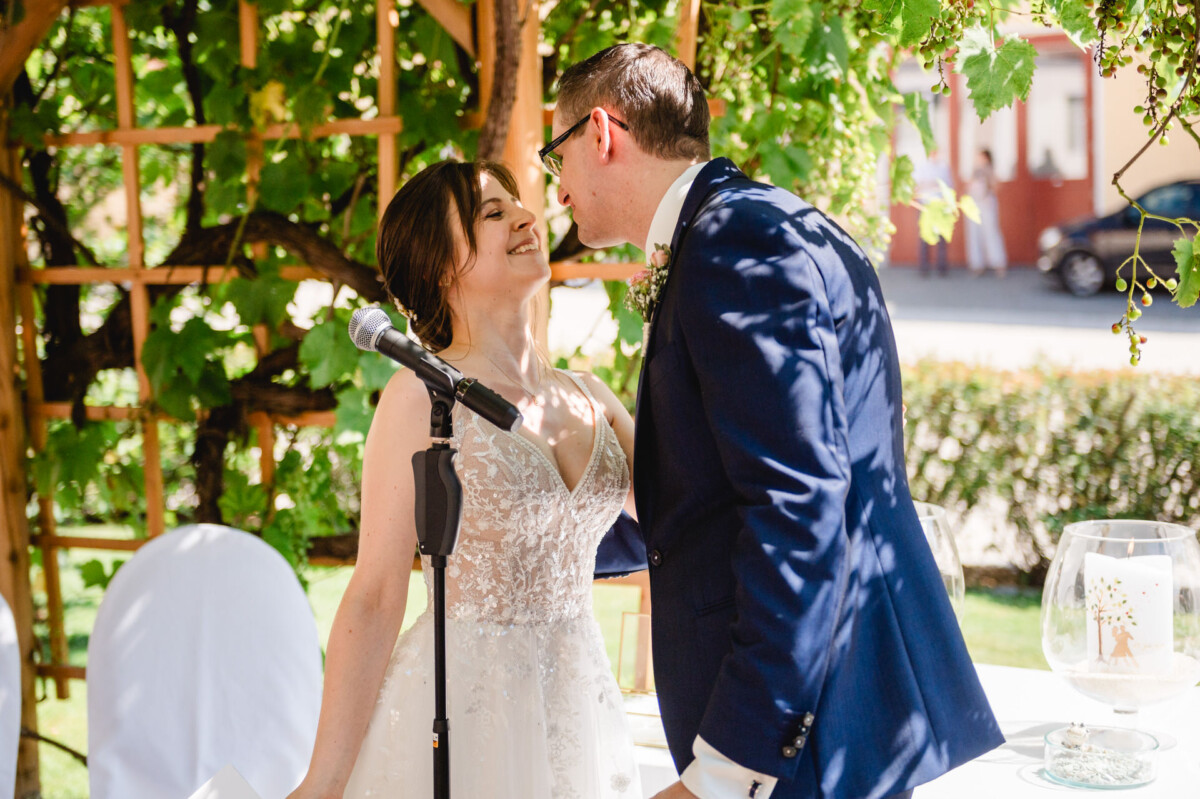 Ein Paar genießt einen Moment bei einem Mikrofonständer während einer Veranstaltung im Freien. Im Hintergrund sieht man viel Grün und ein Holzgitter.