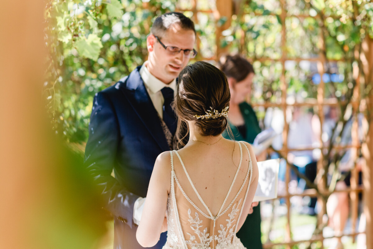 Ein Paar tauscht im Freien unter einer Holzlaube seine Ehegelübde aus. Die Braut trägt ein Spitzenkleid und der Bräutigam einen dunklen Anzug.