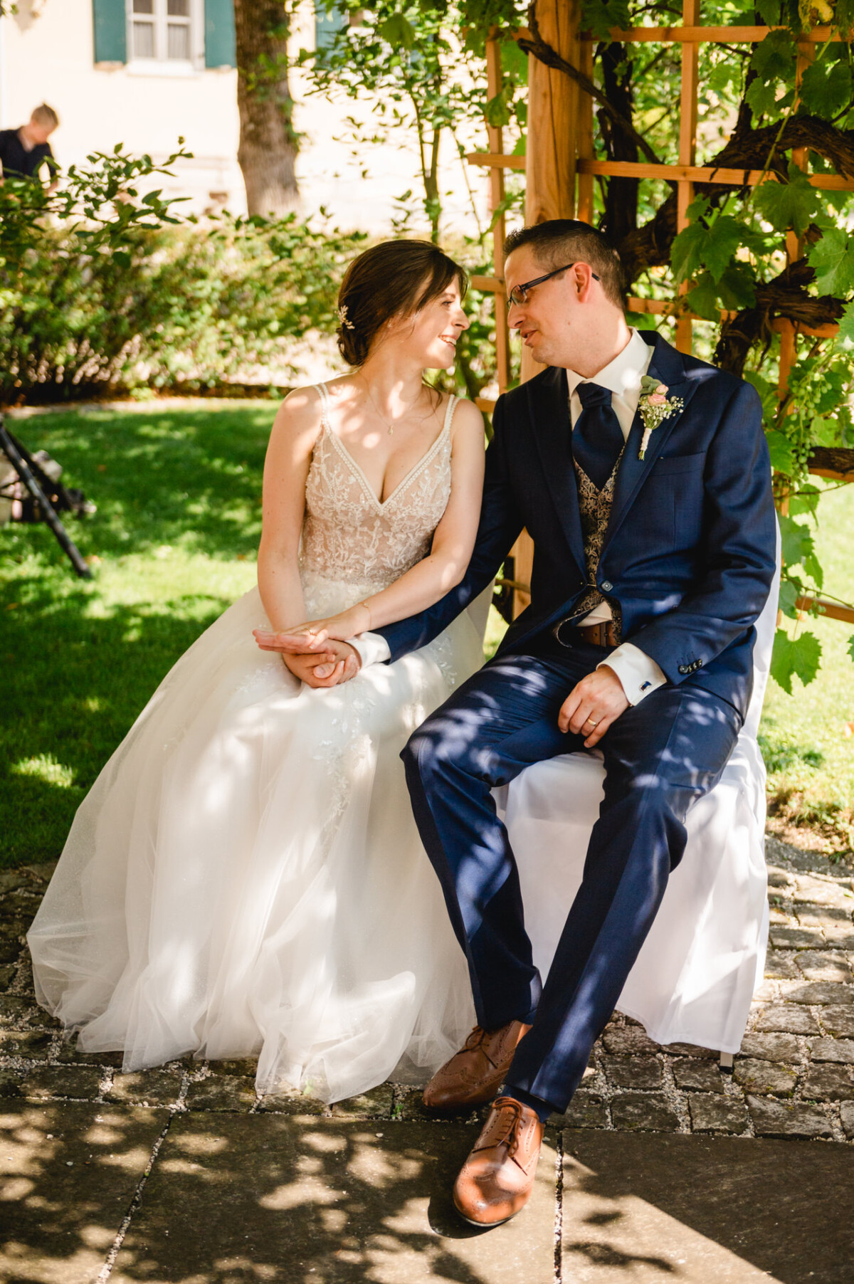 Braut und Bräutigam halten Händchen, während sie im Freien auf einer Bank sitzen, umgeben von Grün und hölzernen Gitterkonstruktionen.