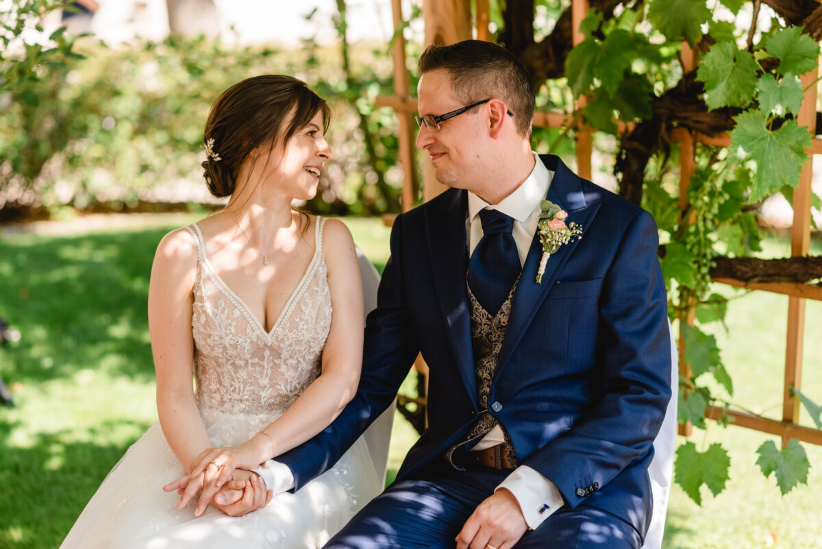 Braut und Bräutigam sitzen draußen unter einer Pergola, halten Händchen und lächeln einander an. Die Braut trägt ein Spitzenkleid und der Bräutigam einen marineblauen Anzug mit Weste und Krawatte.