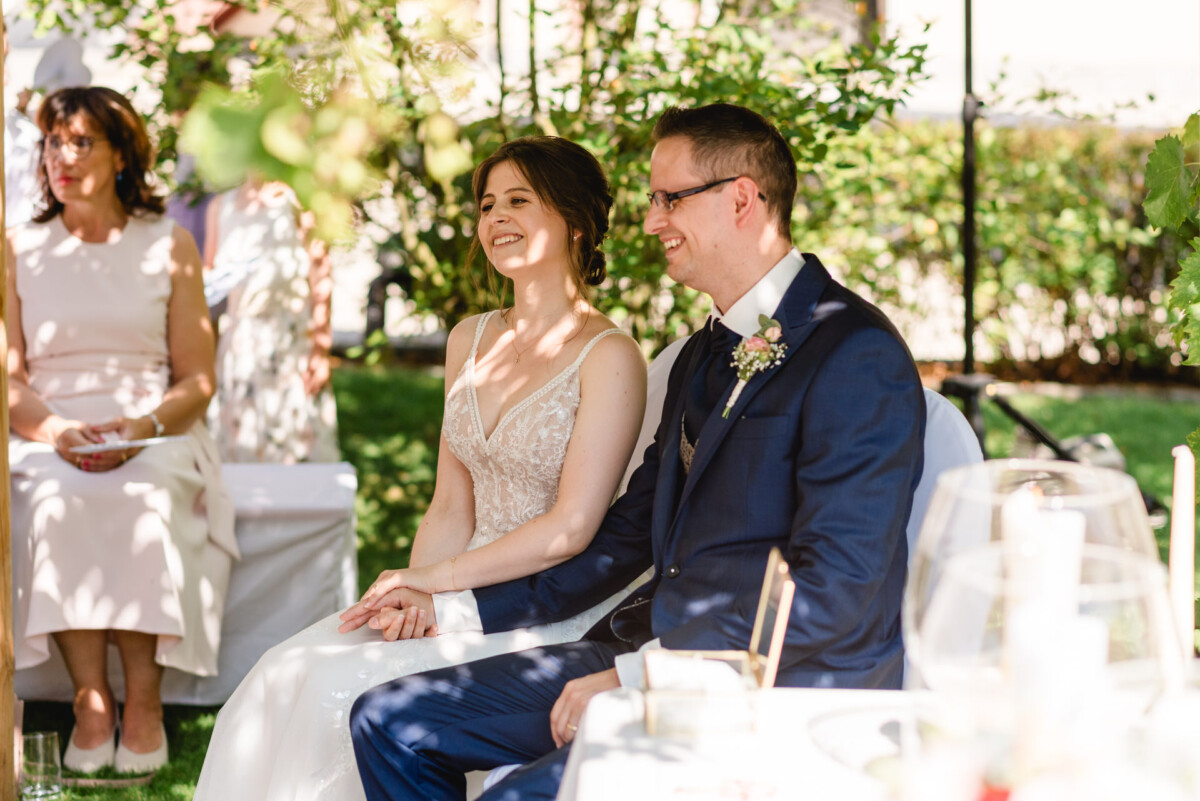Braut und Bräutigam sitzen während einer Hochzeitszeremonie im Freien lächelnd zusammen, umgeben von Gästen.