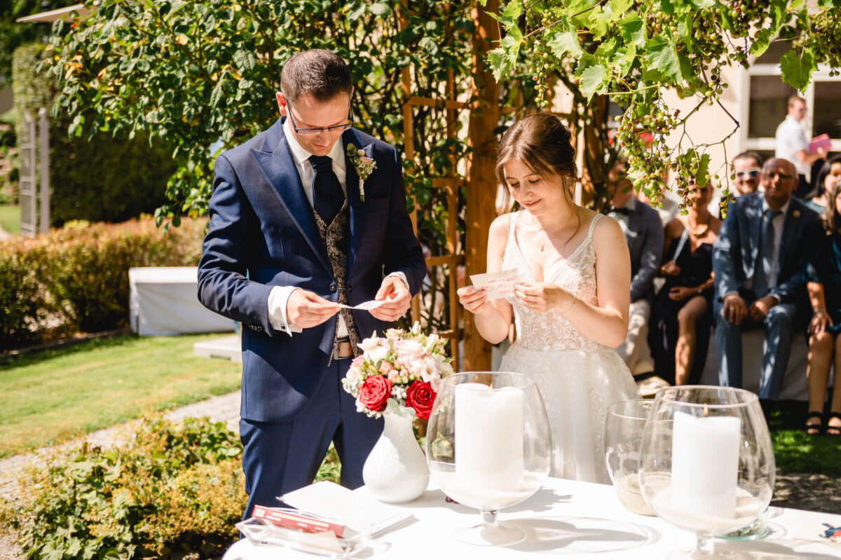 Braut und Bräutigam lesen Notizen bei einer Hochzeitszeremonie im Freien, umgeben von Grün und Gästen.