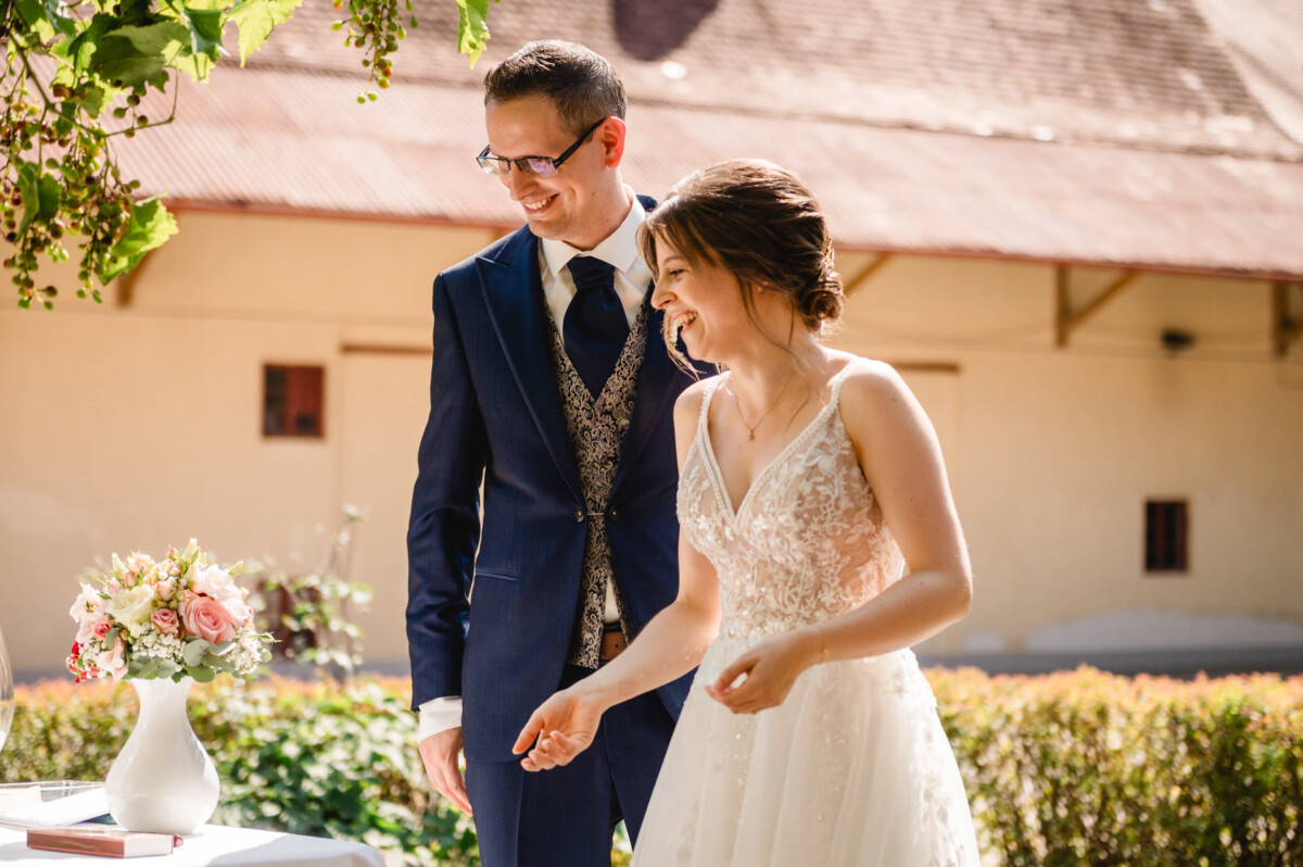 Ein Paar in Abendkleidung, die Frau im weißen Kleid und der Mann im blauen Anzug mit Brille, lächelnd im Freien neben einem Tisch mit Blumen.