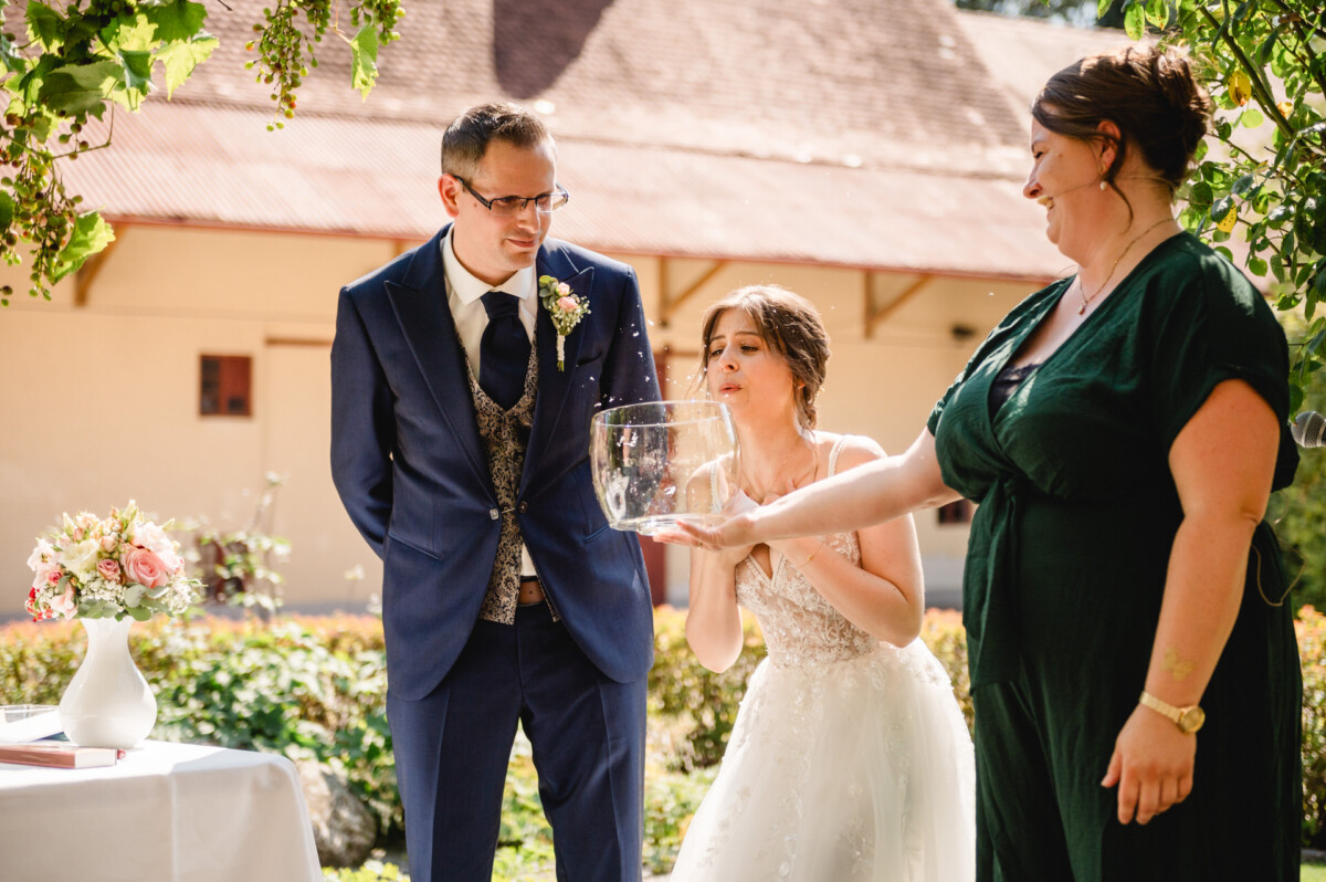 Eine Braut bläst Seifenblasen aus einer Schüssel, die von einer Frau im grünen Kleid gehalten wird, während ein Mann im Anzug daneben steht. Im Freien ist ein Gebäude im Hintergrund zu sehen.