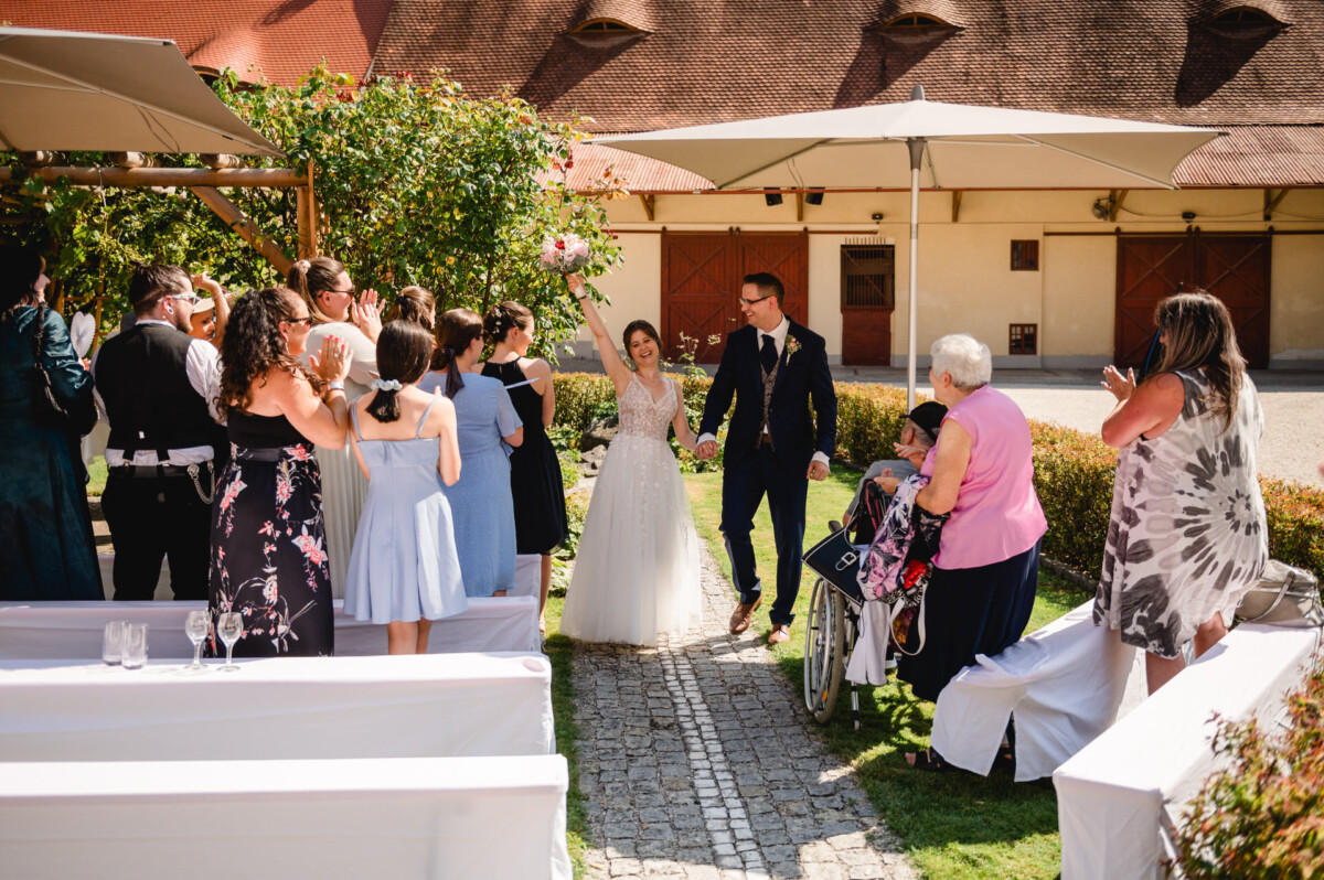 Braut und Bräutigam schreiten im Freien einen Mittelgang entlang, umgeben von applaudierenden Gästen unter Sonnenschirmen.
