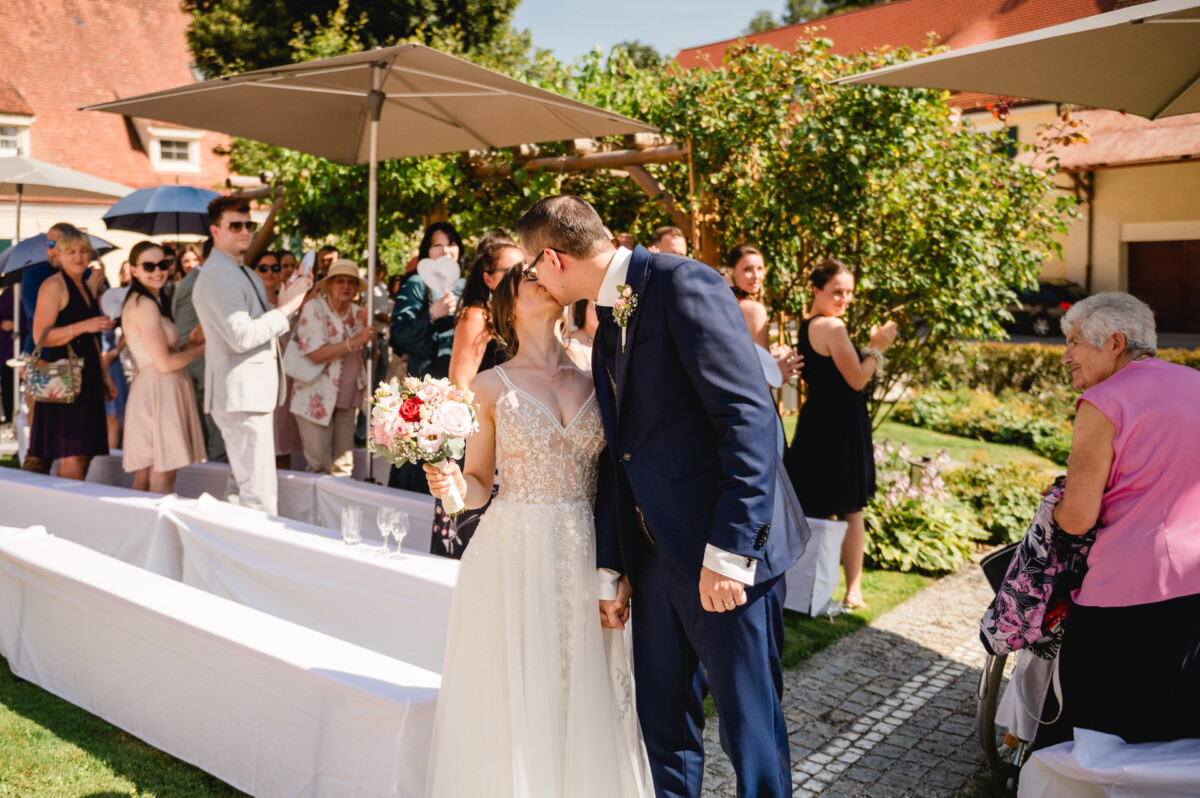 Ein frisch vermähltes Paar küsst sich im Freien, umgeben von Gästen in Abendgarderobe. Die Braut hält einen Blumenstrauß in der Hand und die Szene spielt in einem Garten bei sonnigem Wetter.