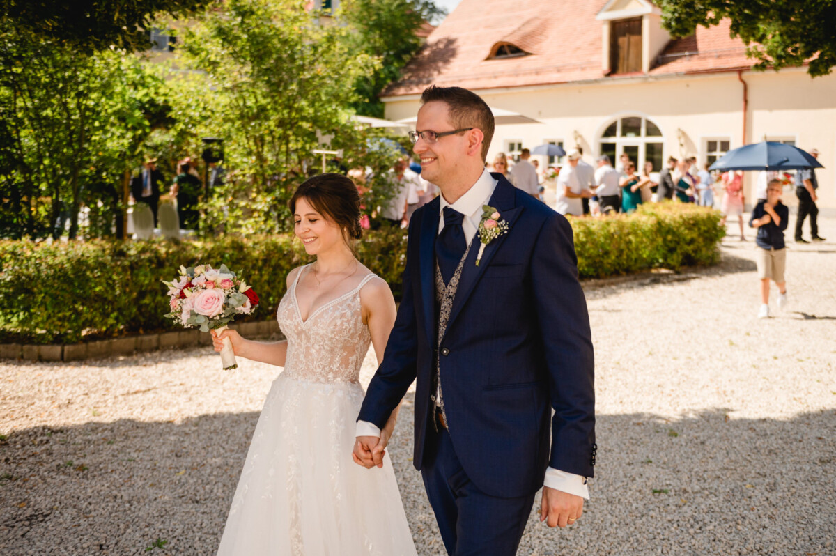 Braut und Bräutigam gehen Hand in Hand im Freien, wobei die Braut einen Blumenstrauß hält. Im Hintergrund ist eine Gruppe von Menschen in der Nähe eines Gebäudes zu sehen.