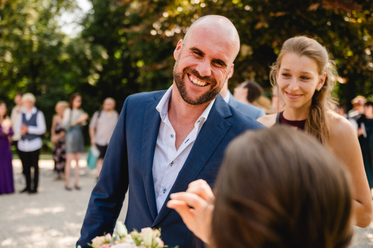 Ein lächelnder Mann im blauen Anzug steht im Freien, neben ihm eine Frau. Im Hintergrund sind Menschen versammelt.