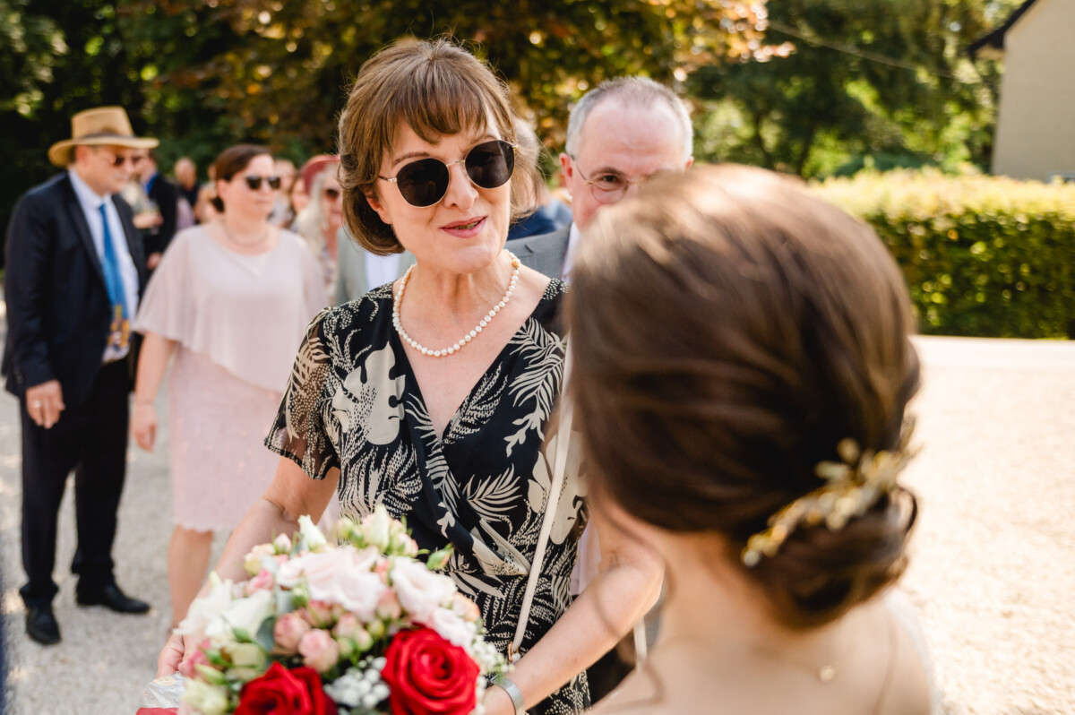 Eine Frau mit Sonnenbrille und geblümtem Kleid hält einen Blumenstrauß in der Hand, während sie sich bei einer Versammlung im Freien mit einer anderen Frau unterhält. Im Hintergrund steht eine Reihe von Menschen.