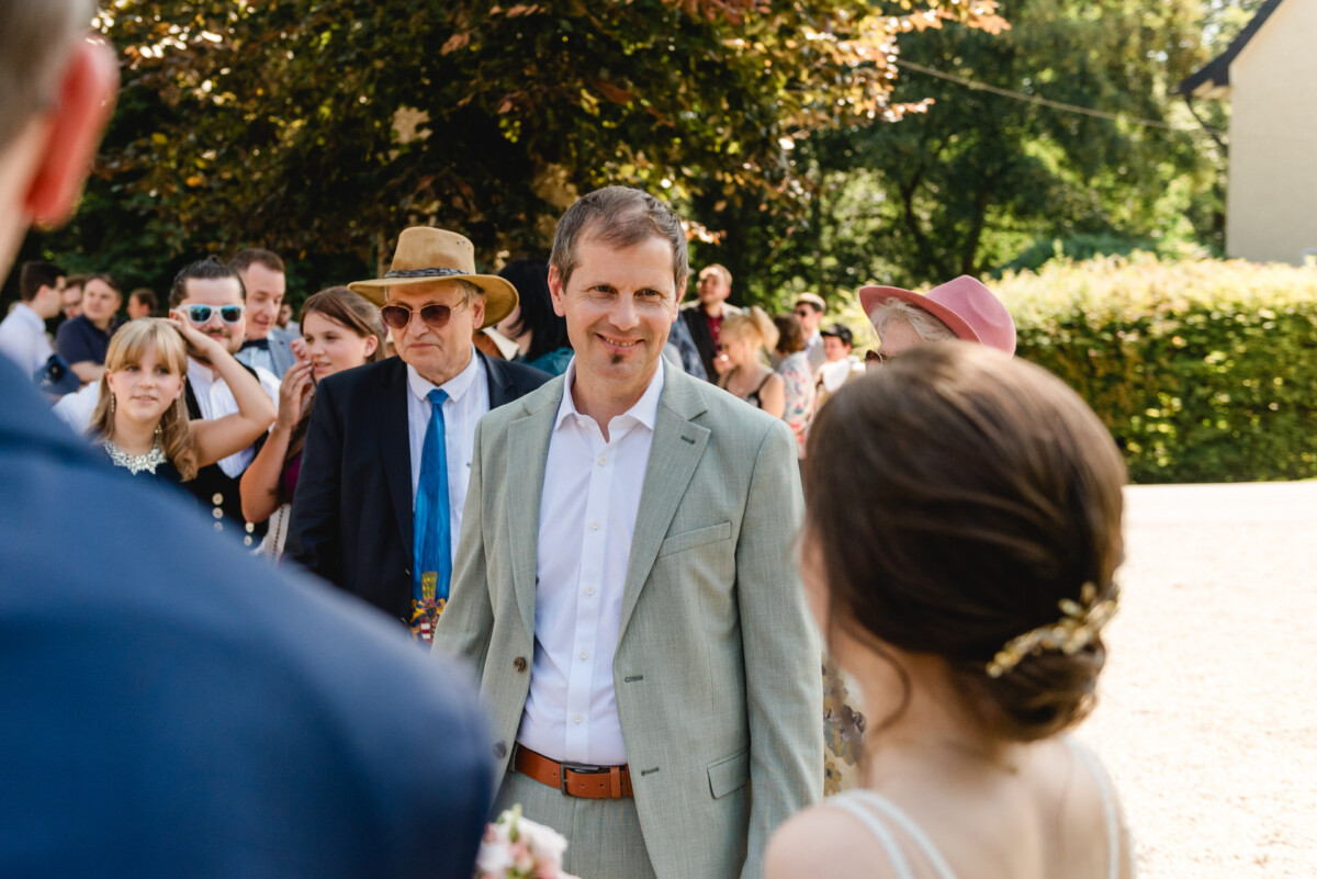 Eine Gruppe formell gekleideter Menschen steht im Freien, im Hintergrund sind Bäume und ein Gebäude zu sehen. Ein Mann in einem hellen Anzug lächelt eine Frau mit Hochsteckfrisur an.