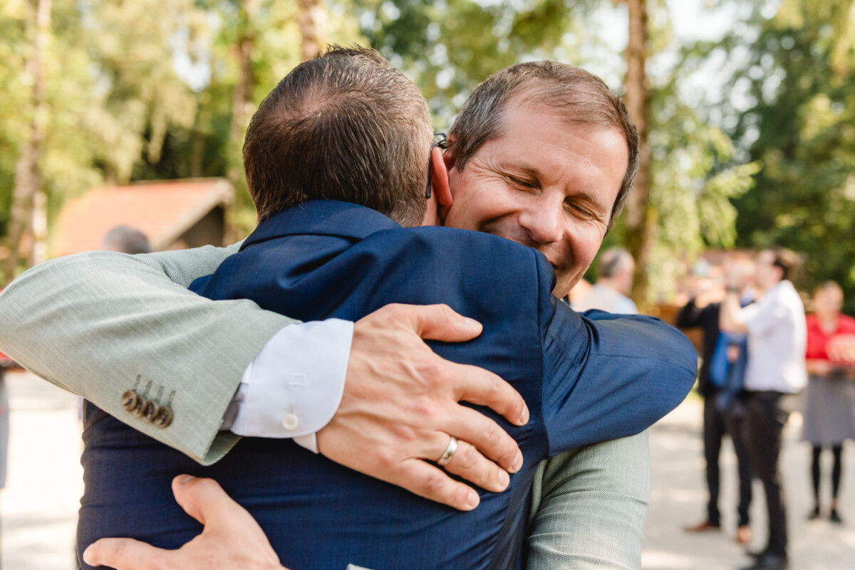 Zwei Männer umarmen sich herzlich im Freien und lächeln beide. Einer trägt einen blauen Anzug, der andere einen hellgrauen. Bäume und Menschen sind im Hintergrund verschwommen.