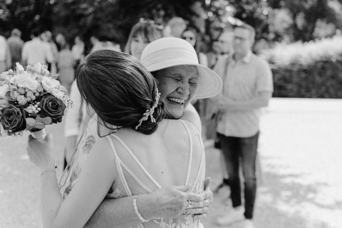 Eine Frau in einem leichten Kleid und Hut umarmt freudig eine andere Frau, die einen Blumenstrauß hält, im Hintergrund sind mehrere Menschen im Freien zu sehen.